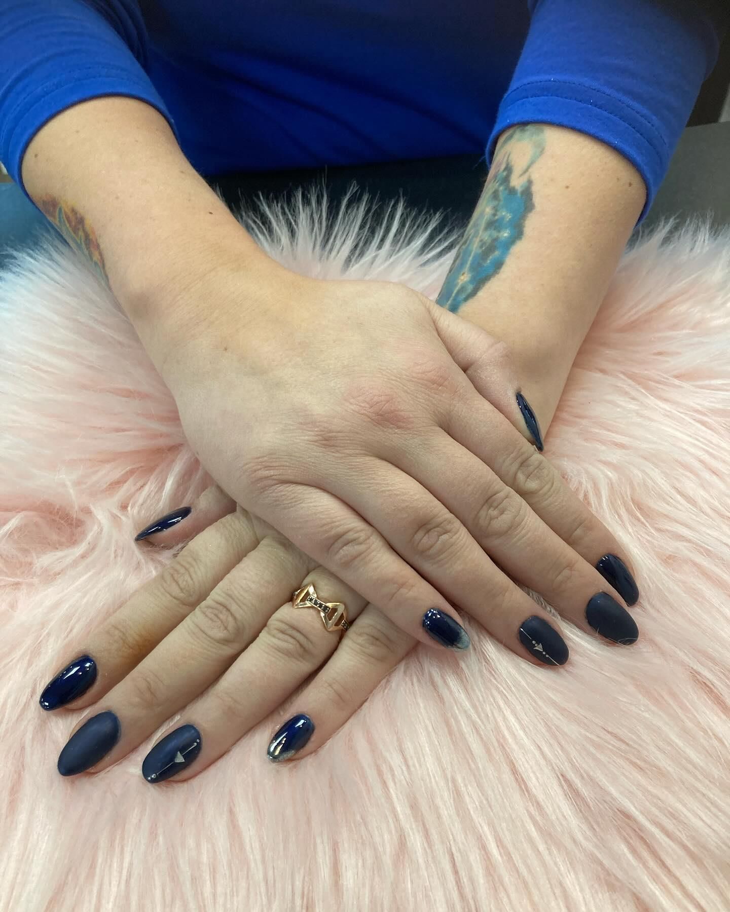 Hands with dark blue matte nails crossed on a pink fur surface, wearing a gold ring.