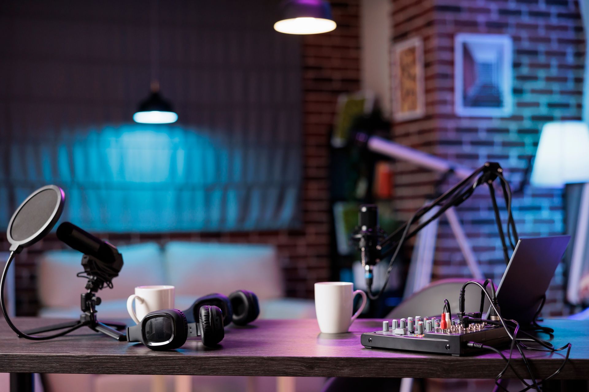 Podcast setup with microphones, headphones, mixing board, and laptop on a desk in a dimly lit room with colorful lighting.