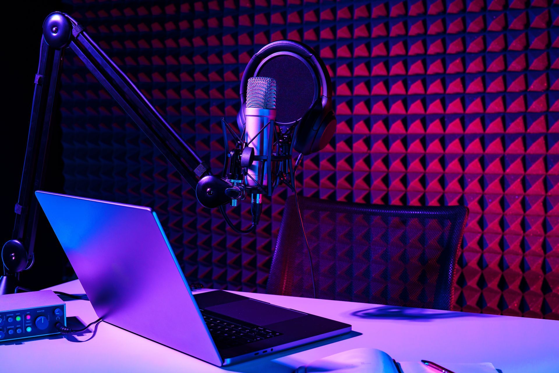 Podcast studio setup with microphone, headphones, laptop, and soundproofing. Illuminated with purple and blue lights.