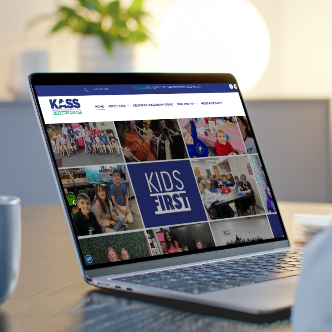 A laptop on a wooden desk displays the KASS (Kentucky Association of School Superintendents) website homepage, featuring a photo grid of students and a 