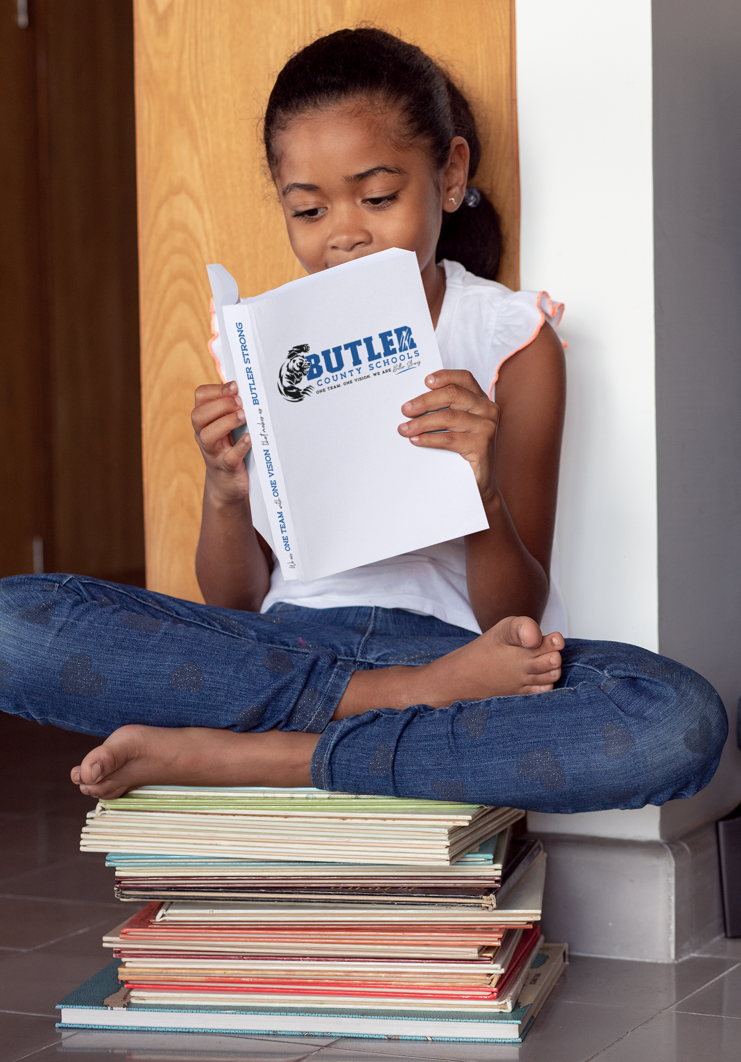 A young girl sits cross-legged with her feet resting on a tall stack of books, reading a Butler County Schools booklet featuring the