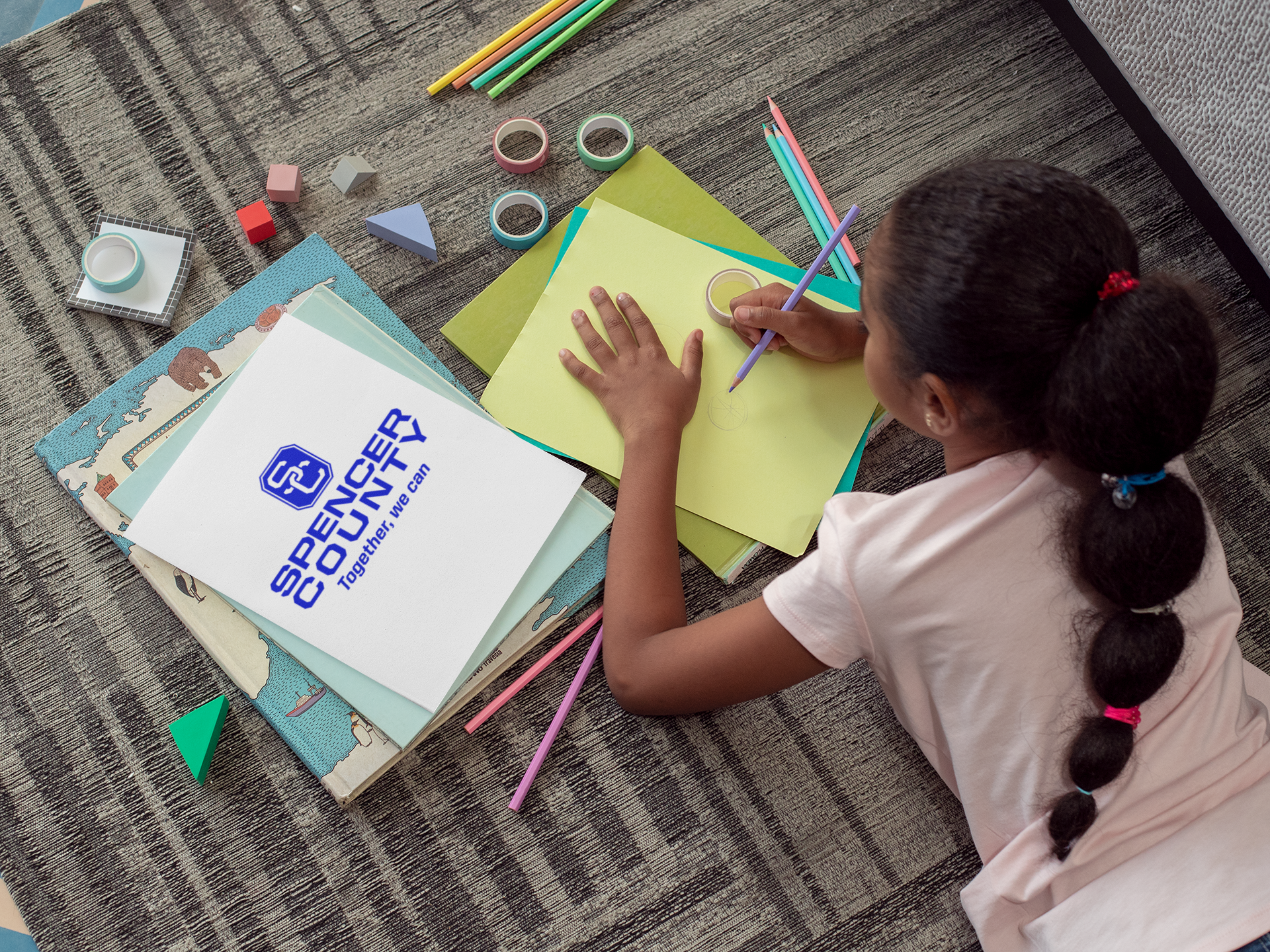An overhead view of a girl with braided hair drawing on colored paper with a pencil, surrounded by craft supplies including washi tape, colored pencils, and a stack of books topped with a