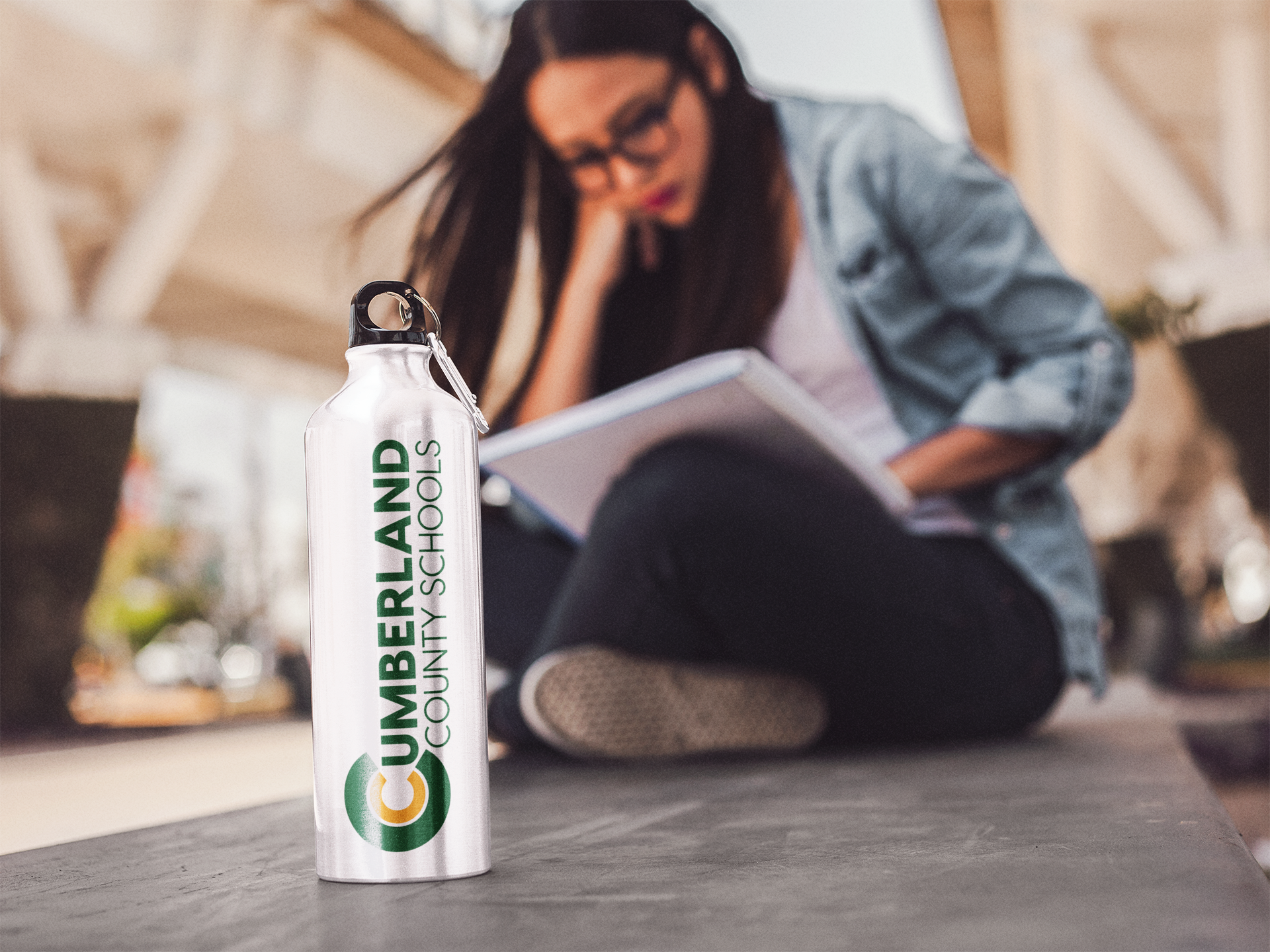 A metal water bottle printed with the Cumberland County Schools logo stands in sharp focus in the foreground, while a young woman sits cross-legged in the blurred background reading a book outdoors.