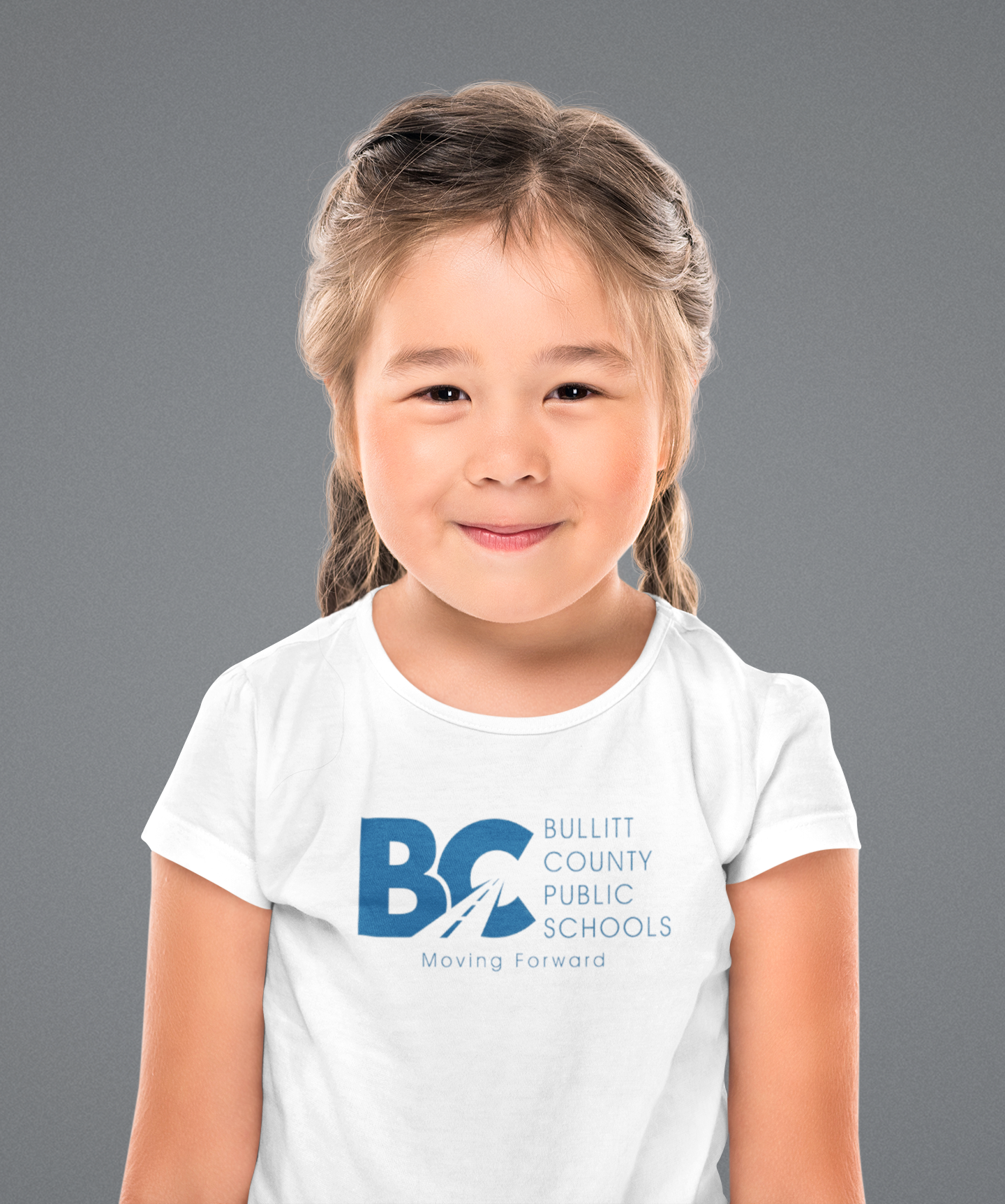 A young girl with braided hair smiles at the camera against a gray background, wearing a white t-shirt printed with the Bullitt County Public Schools