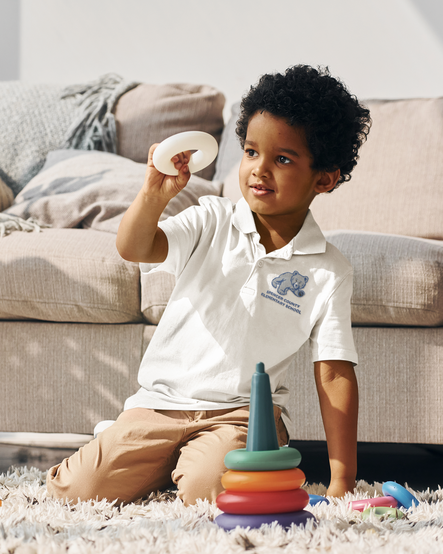 A young boy wearing a white Spencer County Elementary School polo shirt kneels on a rug in a living room, holding up a white ring from a stacking toy while a colorful ring stacker sits in front of him on the floor.