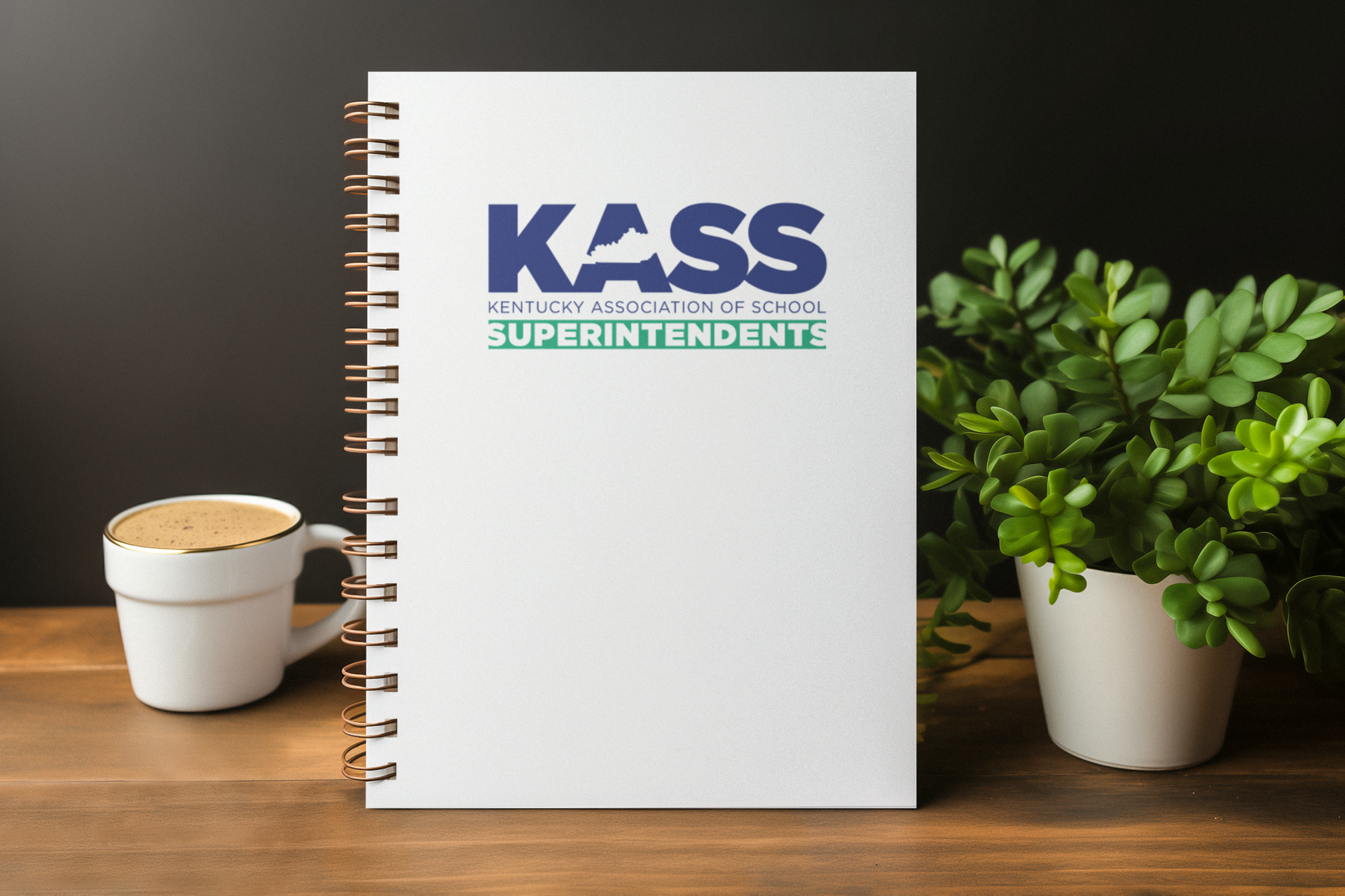 A spiral-bound white notebook with the KASS (Kentucky Association of School Superintendents) logo on the cover, standing upright on a wooden desk between a white latte mug and a small potted jade plant, set against a dark background.