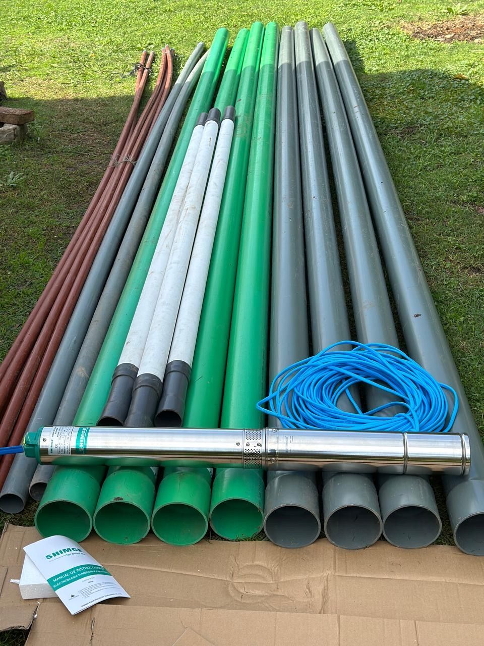 Componentes de una bomba de pozo: varias tuberías (marrón, gris, verde, blanca), bomba sumergible, cable azul enrollado, paquete sobre cartón, al aire libre.