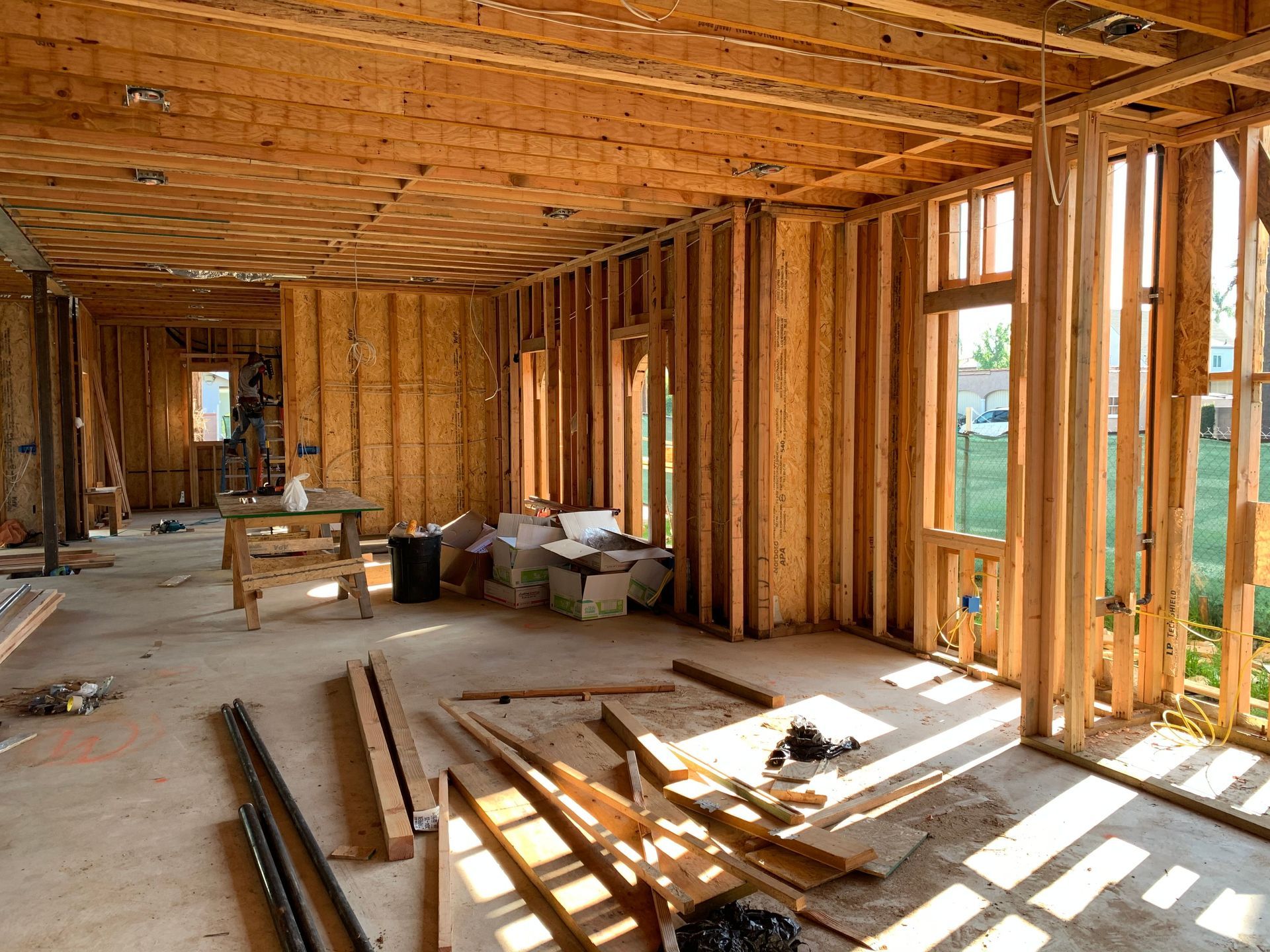 Interior of a wood-framed house under construction with exposed studs, scattered lumber, and sunlight through window openings