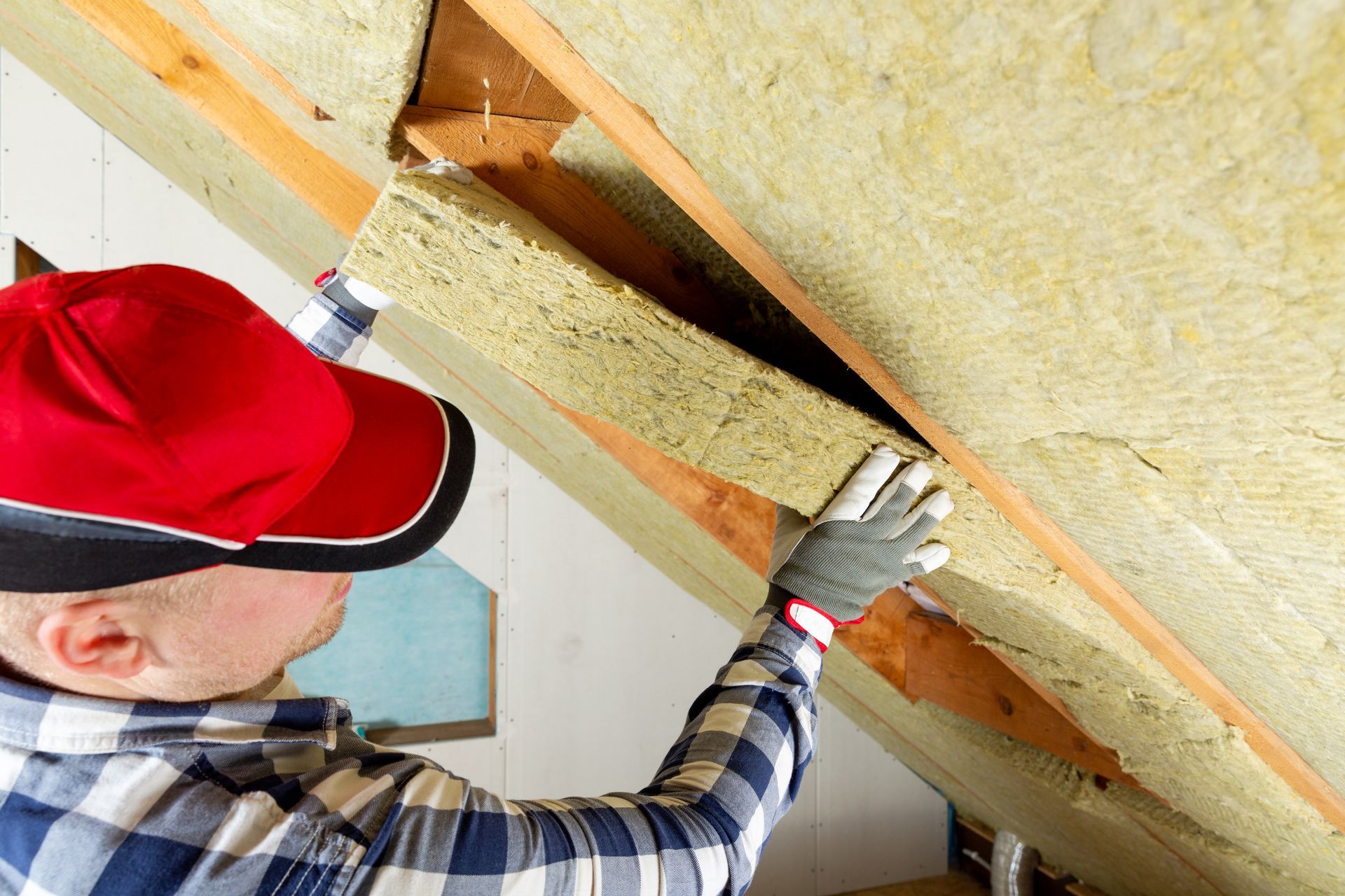 A person wearing a red cap and plaid shirt installs yellow insulation panels between wooden roof rafters.