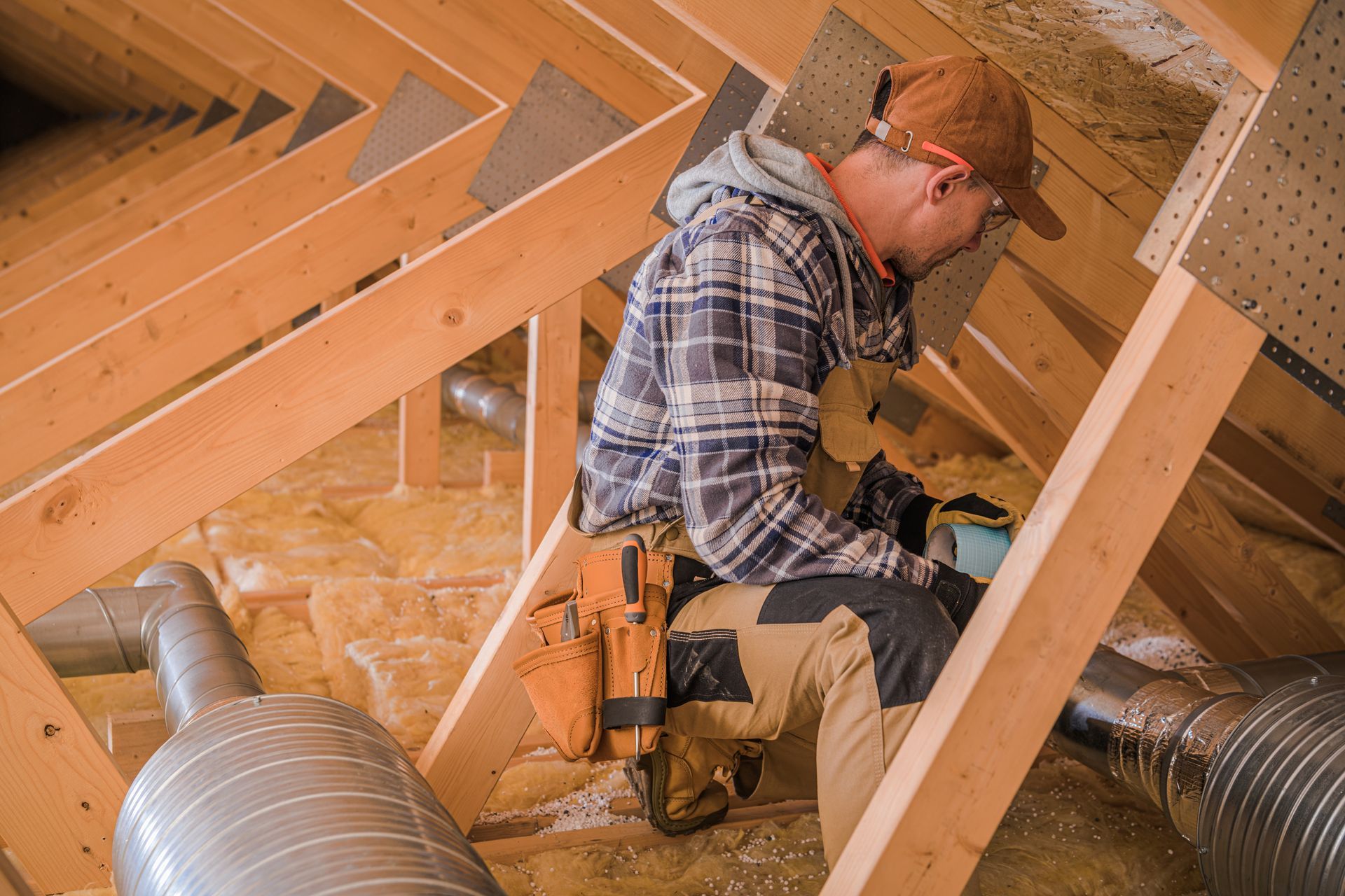 Man installing insulation in an attic, wearing a hat, glasses, and tool belt.
