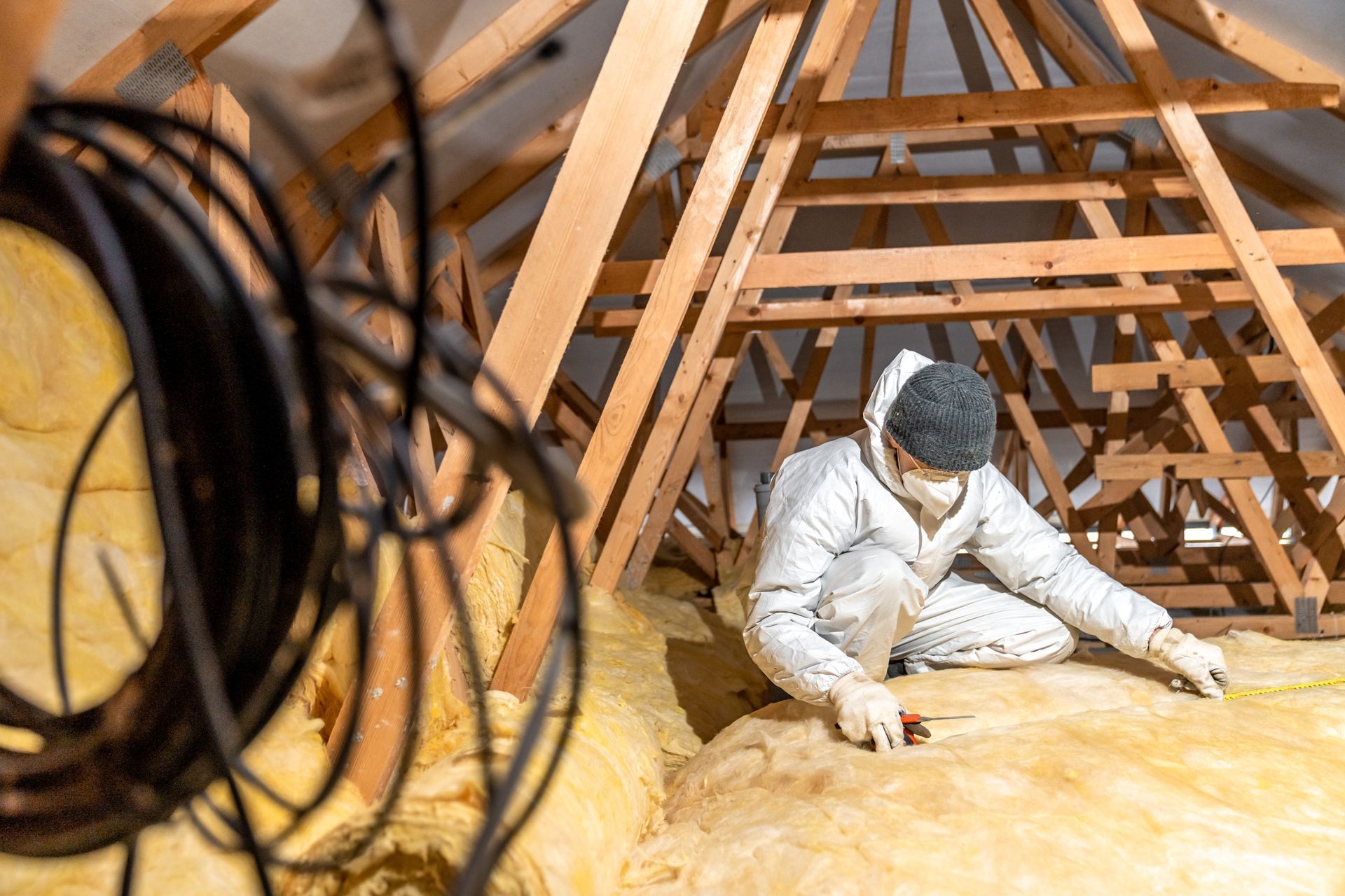 Worker installing insulation in an attic, surrounded by wooden rafters and exposed wiring.