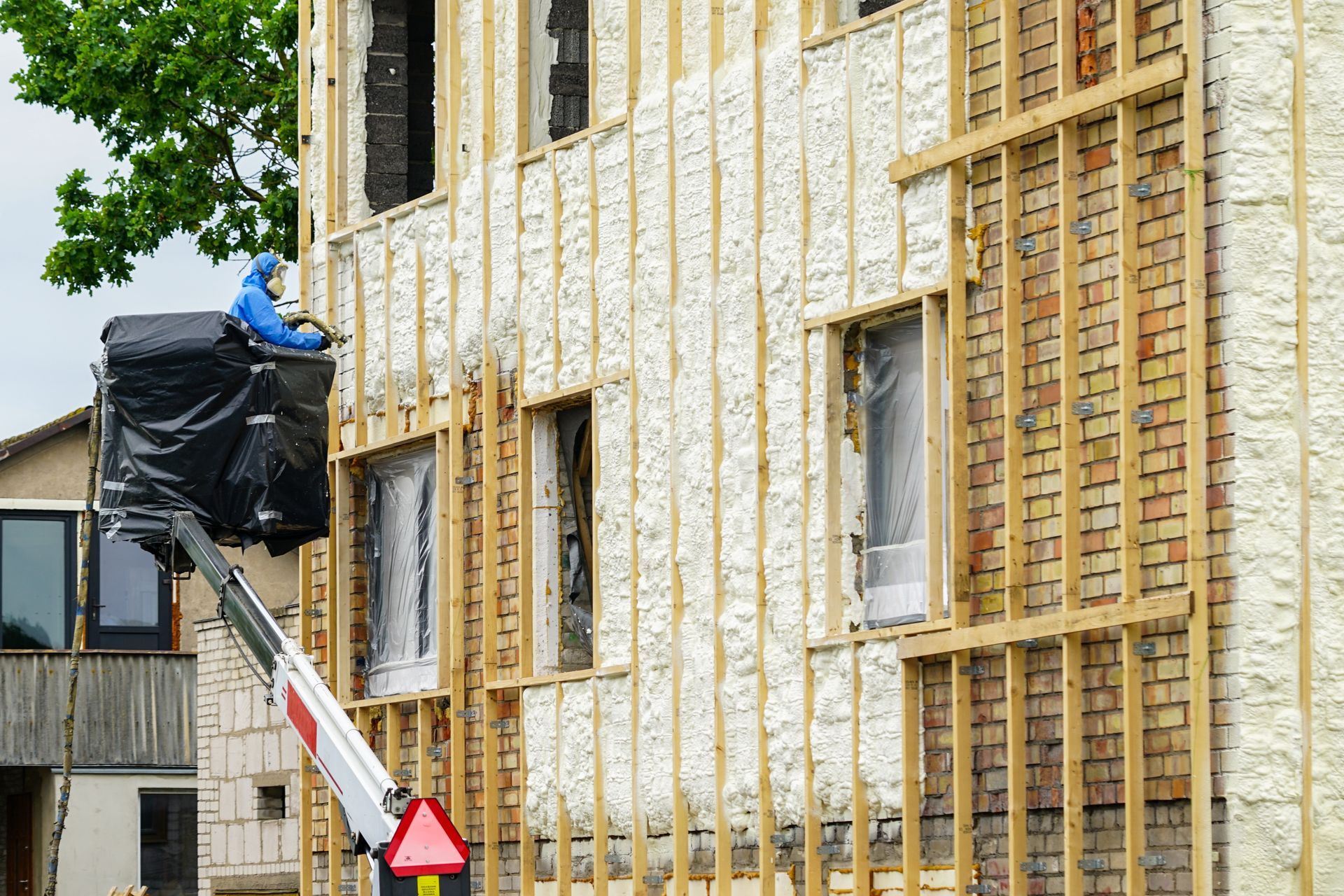 Worker in lift sprays insulation on building exterior. Brick, wood frame, windows.