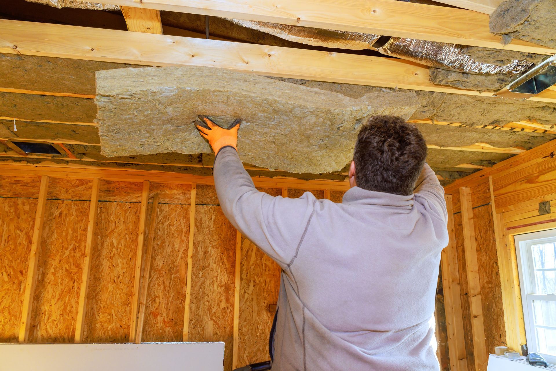 Person installing insulation in an unfinished attic with exposed wooden framing.