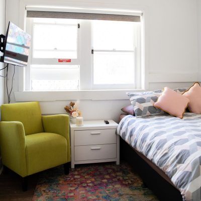 Bedroom with green armchair, bed with pink pillows, white nightstand, and TV mounted on the wall near a window.