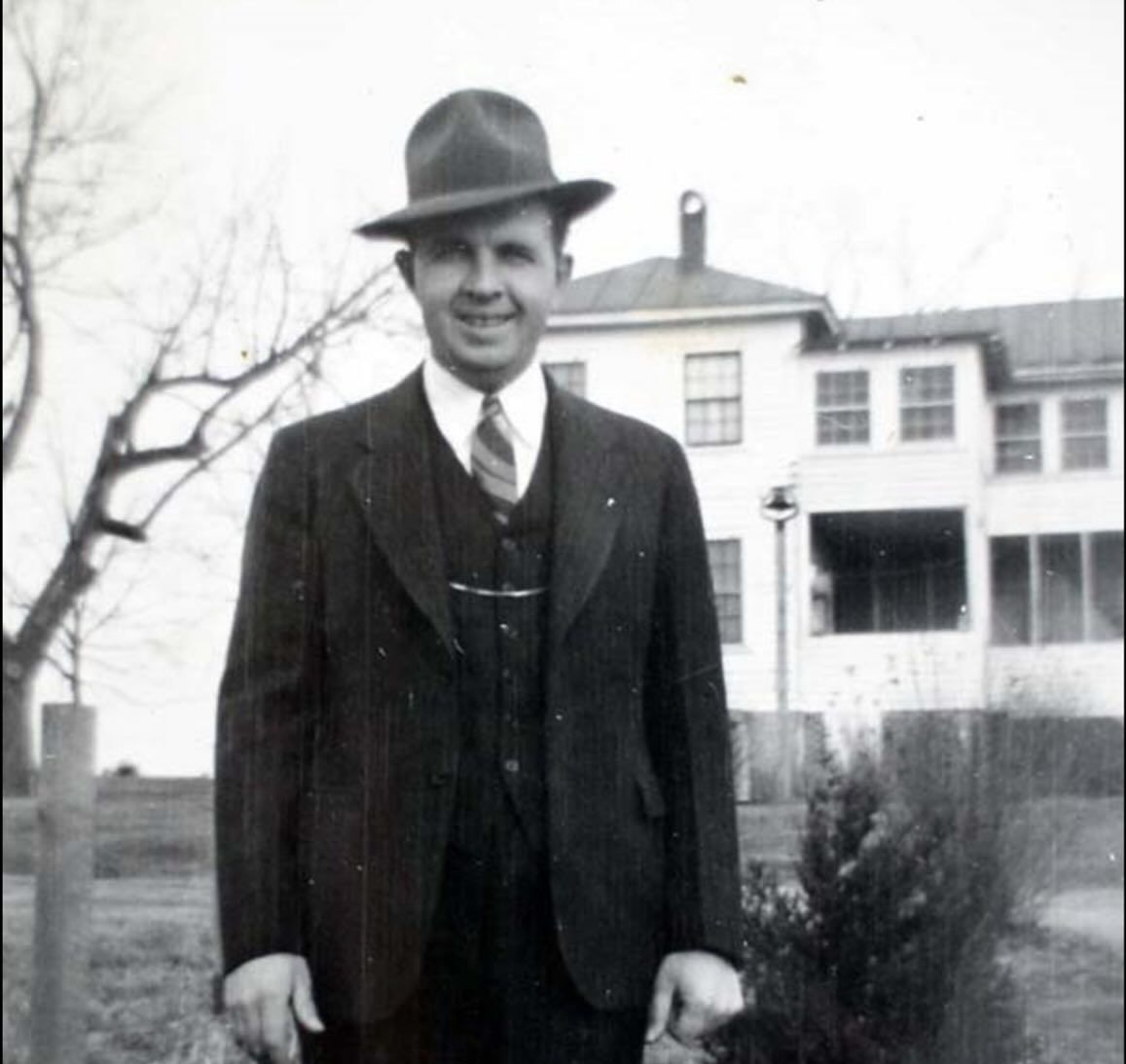 A person wearing a fedora and a suit with a vest stands in front of a white two-story house with a large porch.