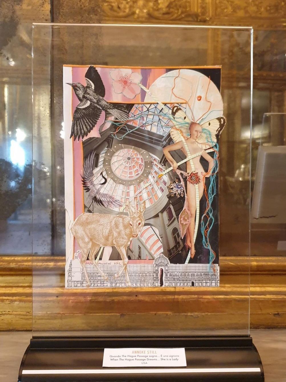 Collagekunstwerk: vogel, naaktfiguur, geit en klokachtig object, achter een transparante acrylvitrine, op een displaystandaard.