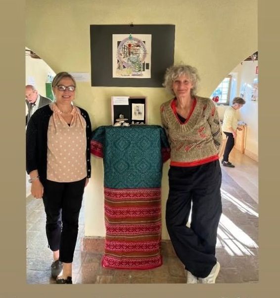 Twee vrouwen staan naast een kunsttentoonstelling met textiel en ingelijste kunstwerken. Ze glimlachen. De tentoonstelling bevindt zich in een gang.