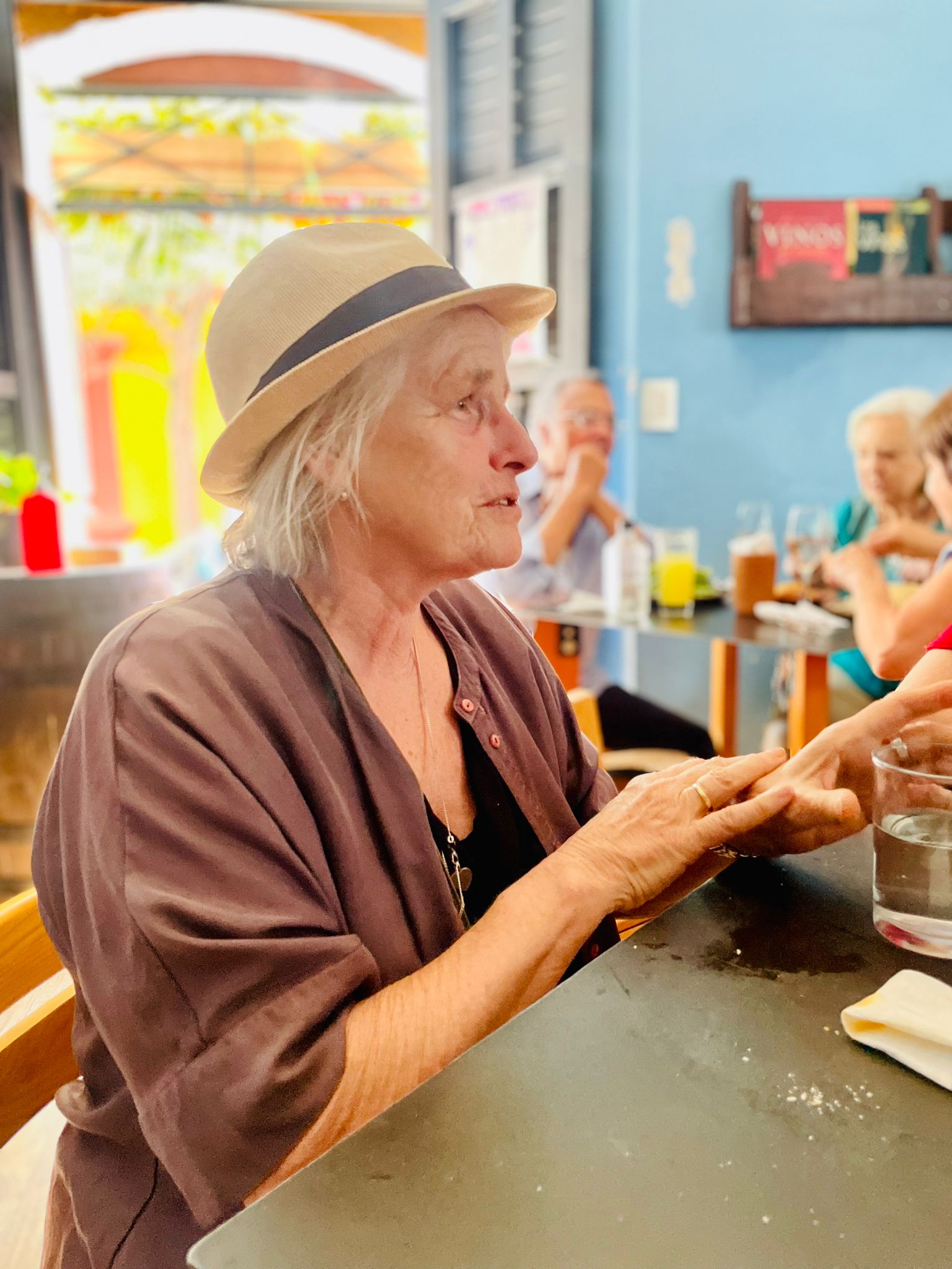 Photo Credits Helen Preston Hartmann Vrouw met hoed in een café, gebarend met haar handen, zittend aan een tafel. Blauwe muur en andere gasten op de achtergrond.