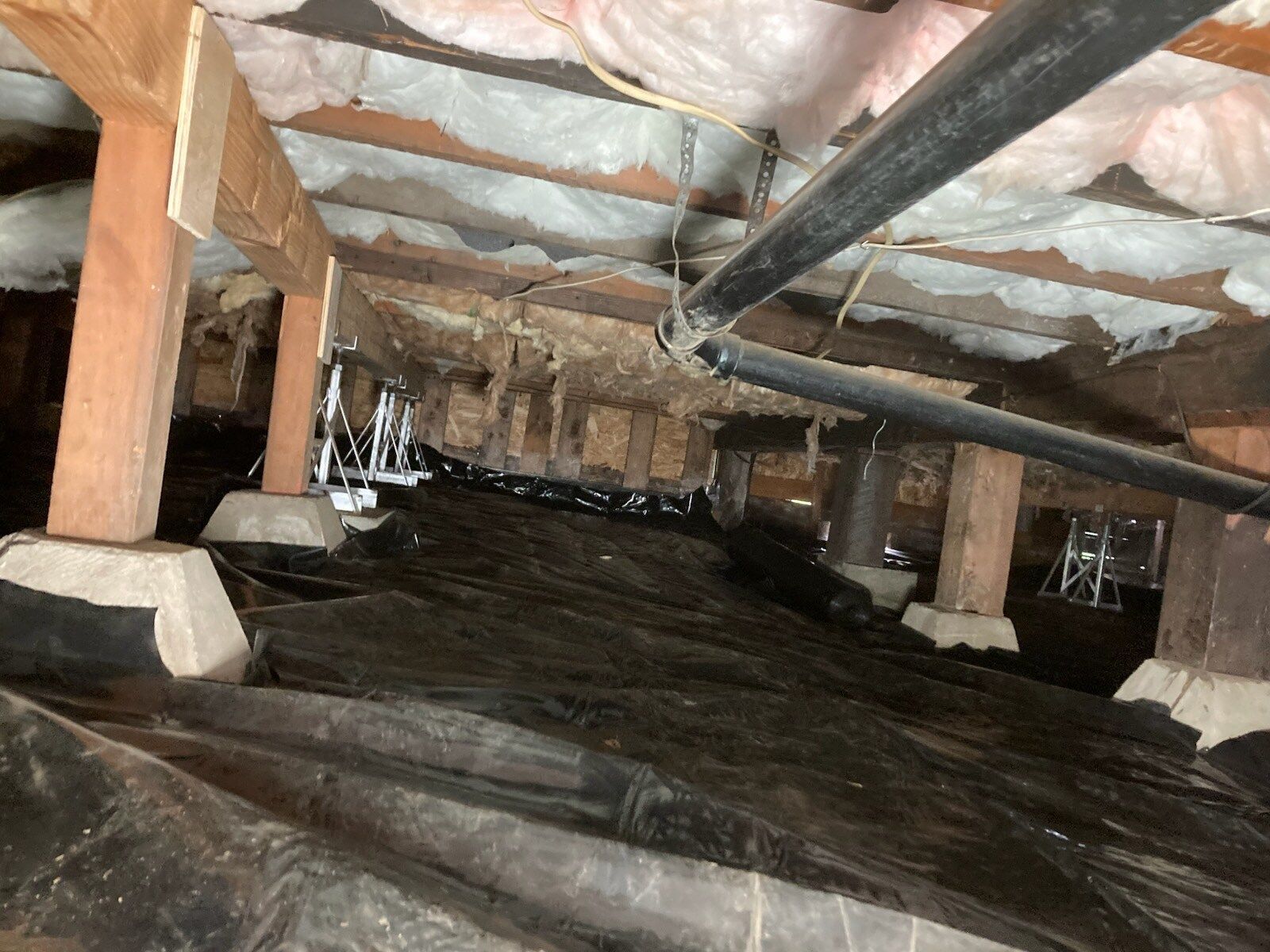 View from under a house with a black vapor barrier, insulation, pipes, and support posts.