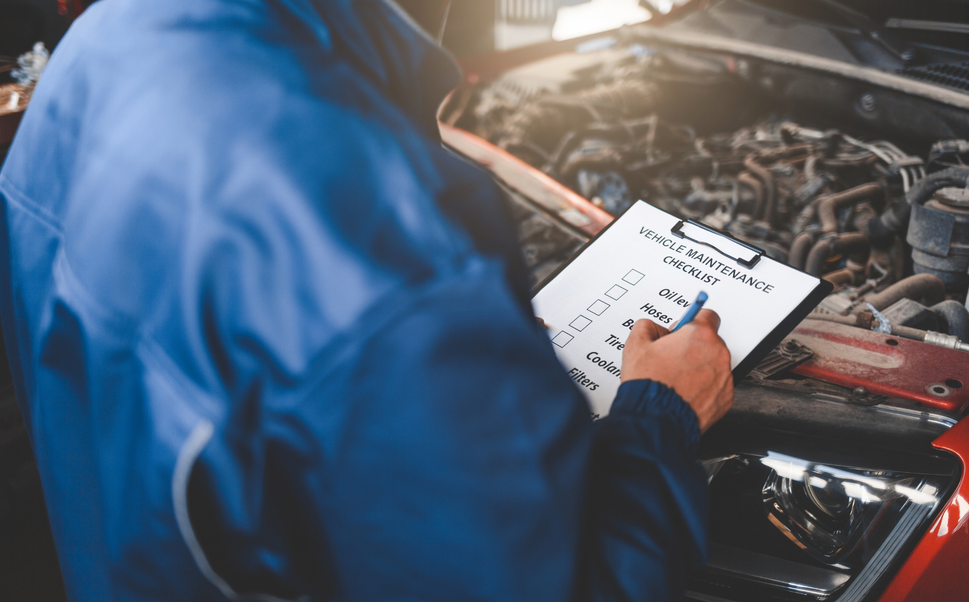 Mechanic in blue coveralls inspecting a car engine on a clipboard