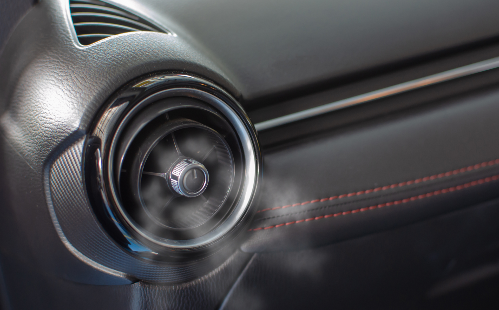 Close-up of a car dashboard air vent with black textured trim and red stitching.