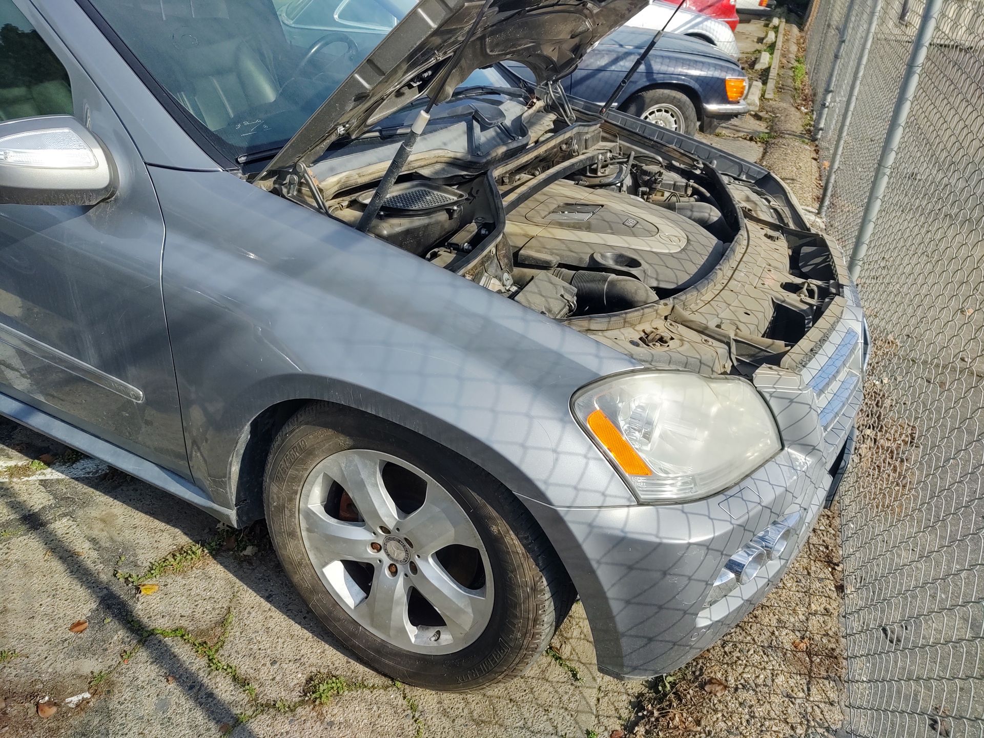 Gray SUV with hood open, exposing engine, parked on a gravel lot by a chain-link fence