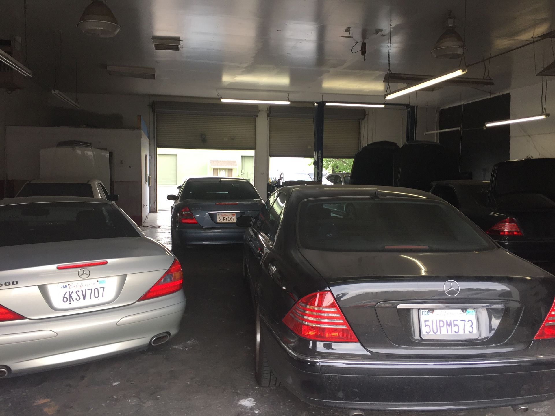 Cars parked inside a dim auto shop with garage doors open to daylight.