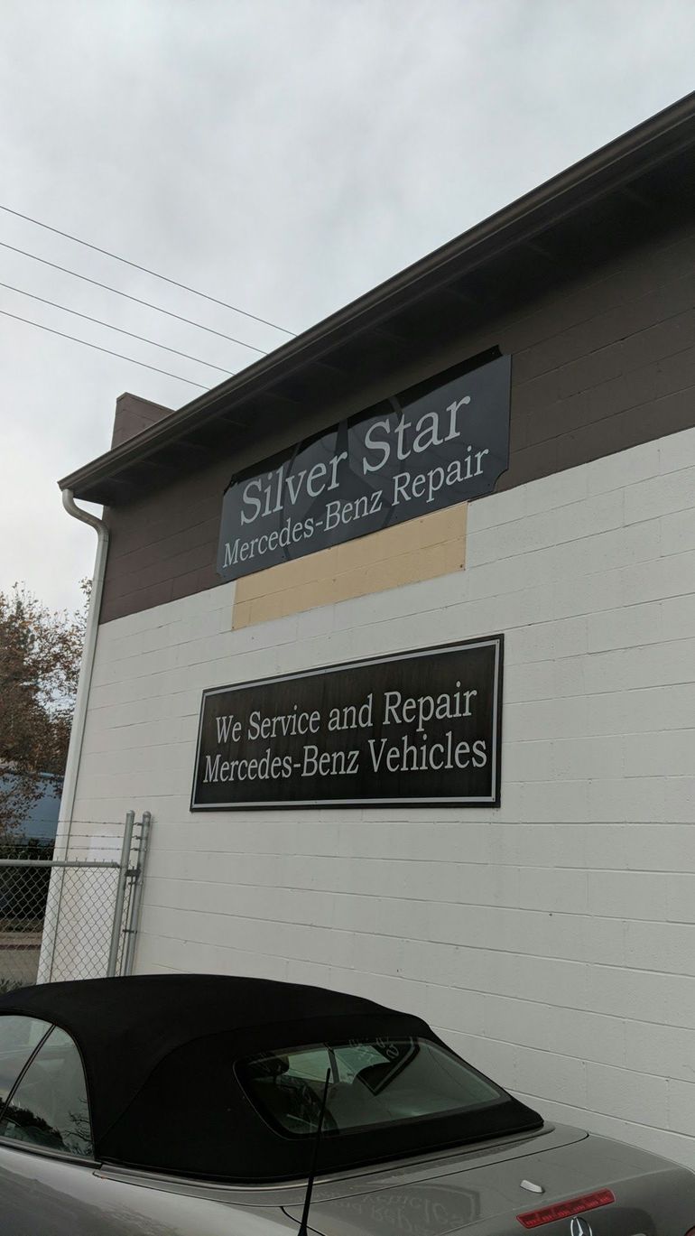 Auto repair shop exterior with black and white signs and a parked gray car under a cloudy sky