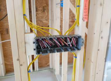 Electrical junction box with exposed wires, mounted on wooden studs in a construction setting.