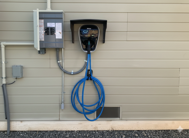 Electric vehicle charger installed on a beige house wall, with a breaker box and blue charging cable.