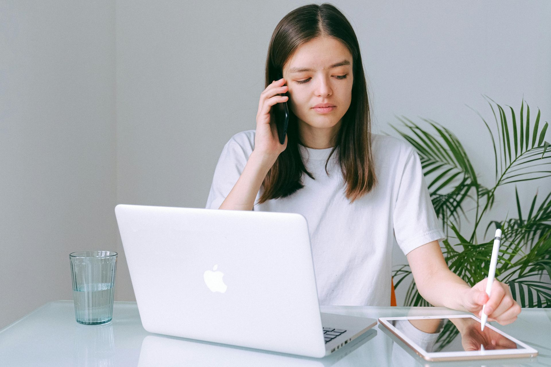 Een vrouw zit aan een bureau met een laptop en een tablet terwijl ze op een mobiele telefoon praat.