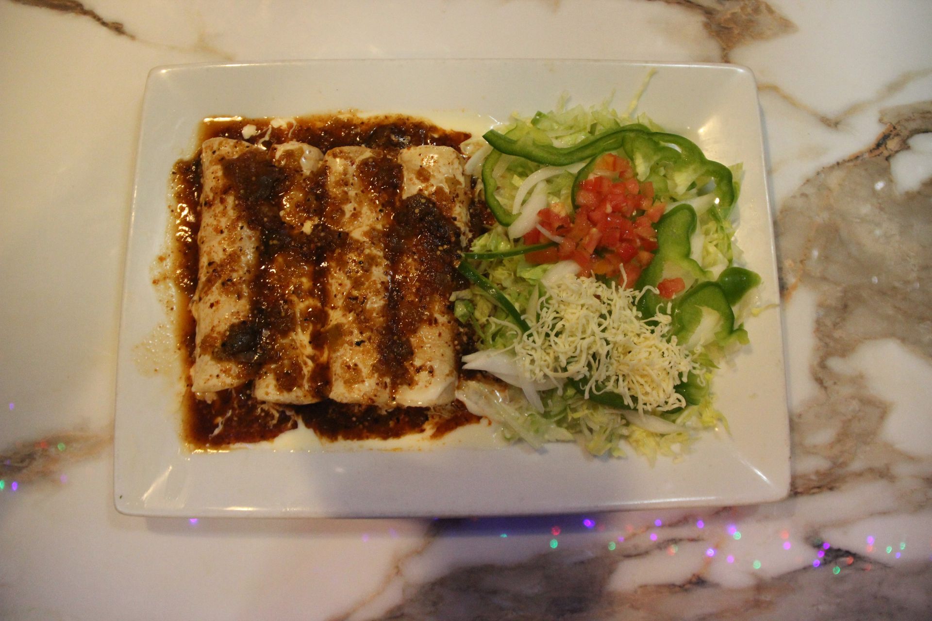 Plated grilled entrée with brown sauce beside mixed greens, shredded cheese, and diced tomatoes on a marble table