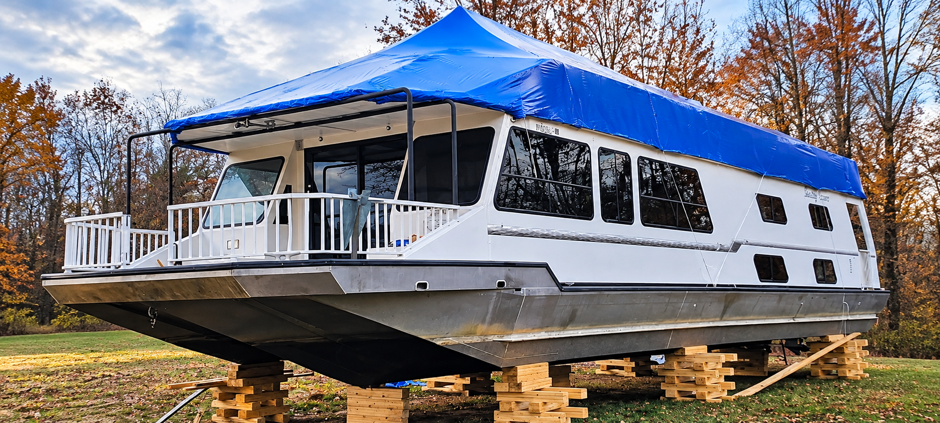 Large white houseboat with a blue tarp roof on wooden blocks in a yard, viewed from the front-left.