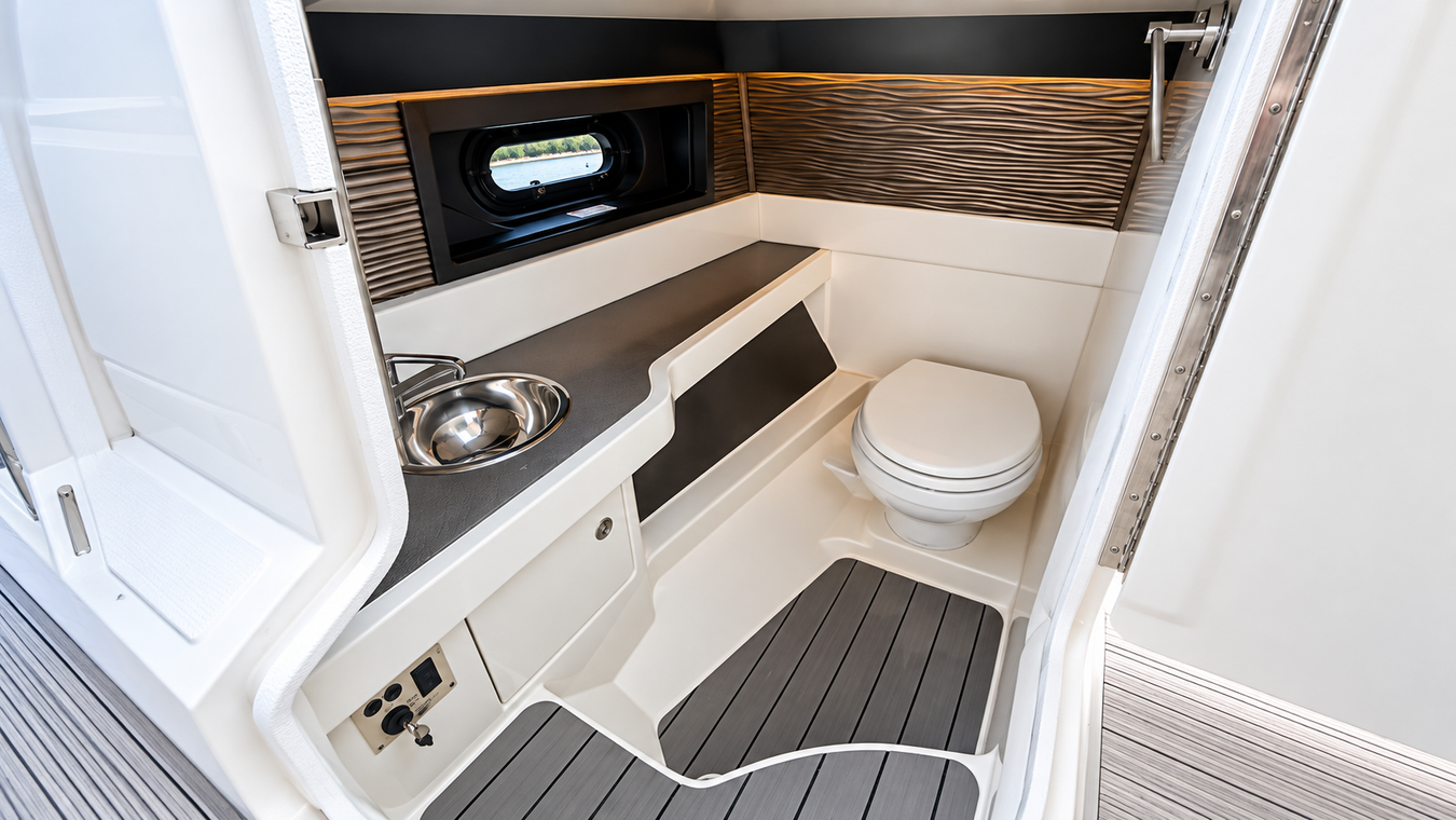 Small modern boat or RV bathroom with toilet, sink, and gray floor mat, finished in white and wood tones