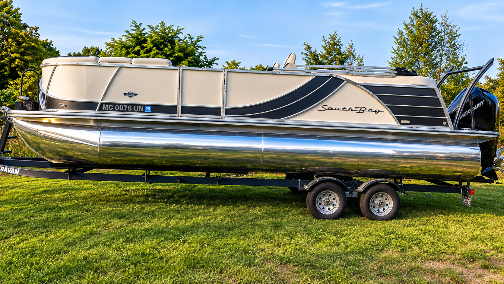 Silver pontoon boat on a trailer parked on grass under a blue sky.
