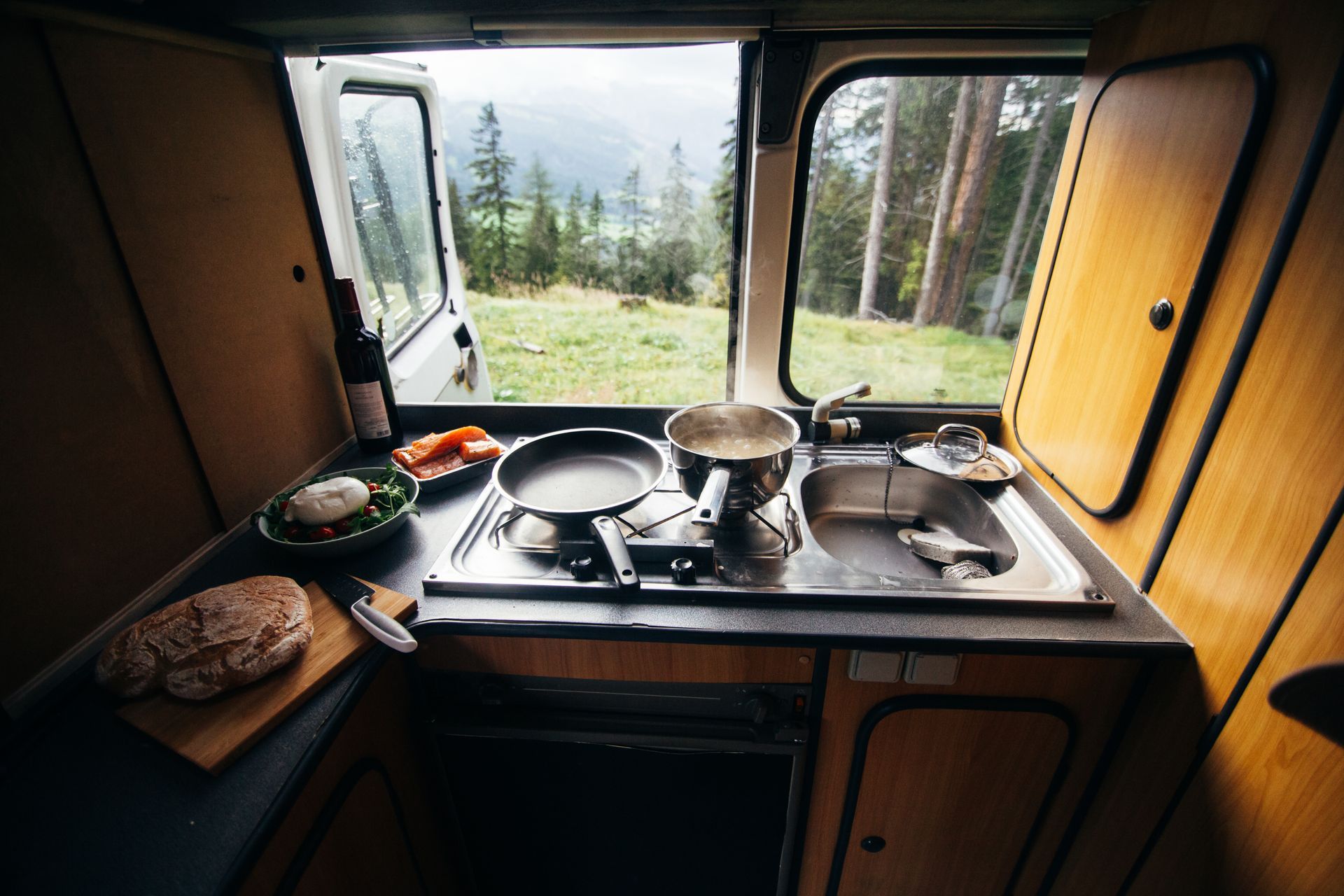 Compact camper kitchen with sink, stove, pan, dishes, and forest view through open windows