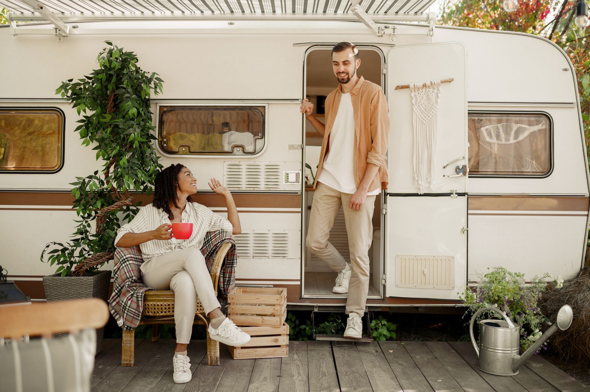 Couple by a vintage camper, one seated with a cup and one standing in the doorway on a porch-like deck