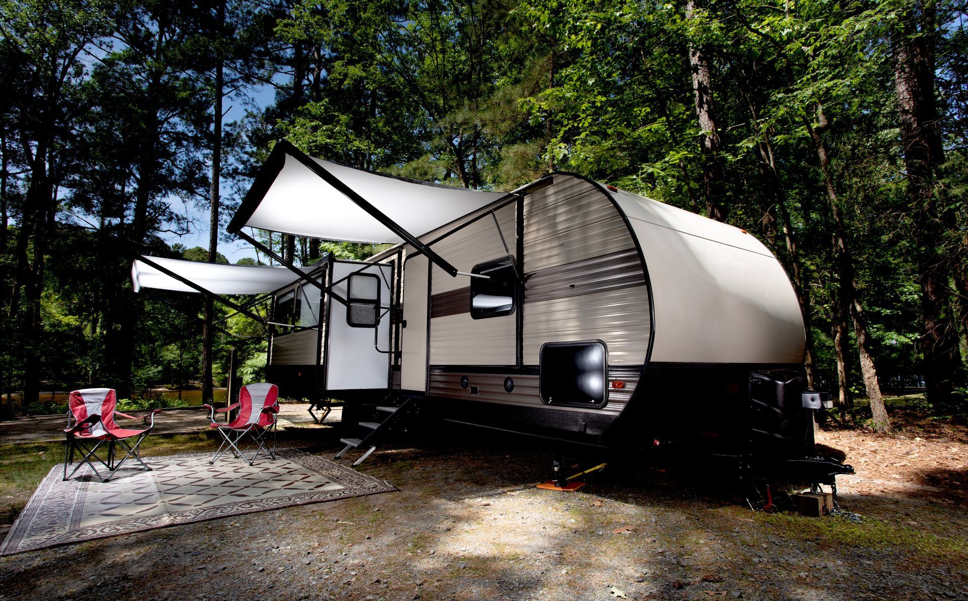 Modern white camper trailer in a wooded campsite with awning and pink chairs
