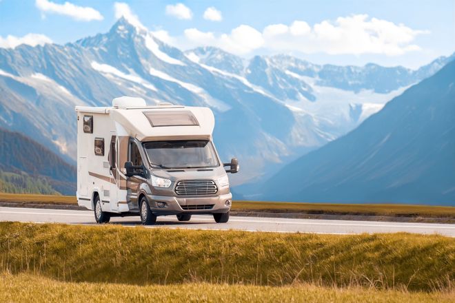 White RV driving on a mountain road with snowy peaks and grassy fields in the background