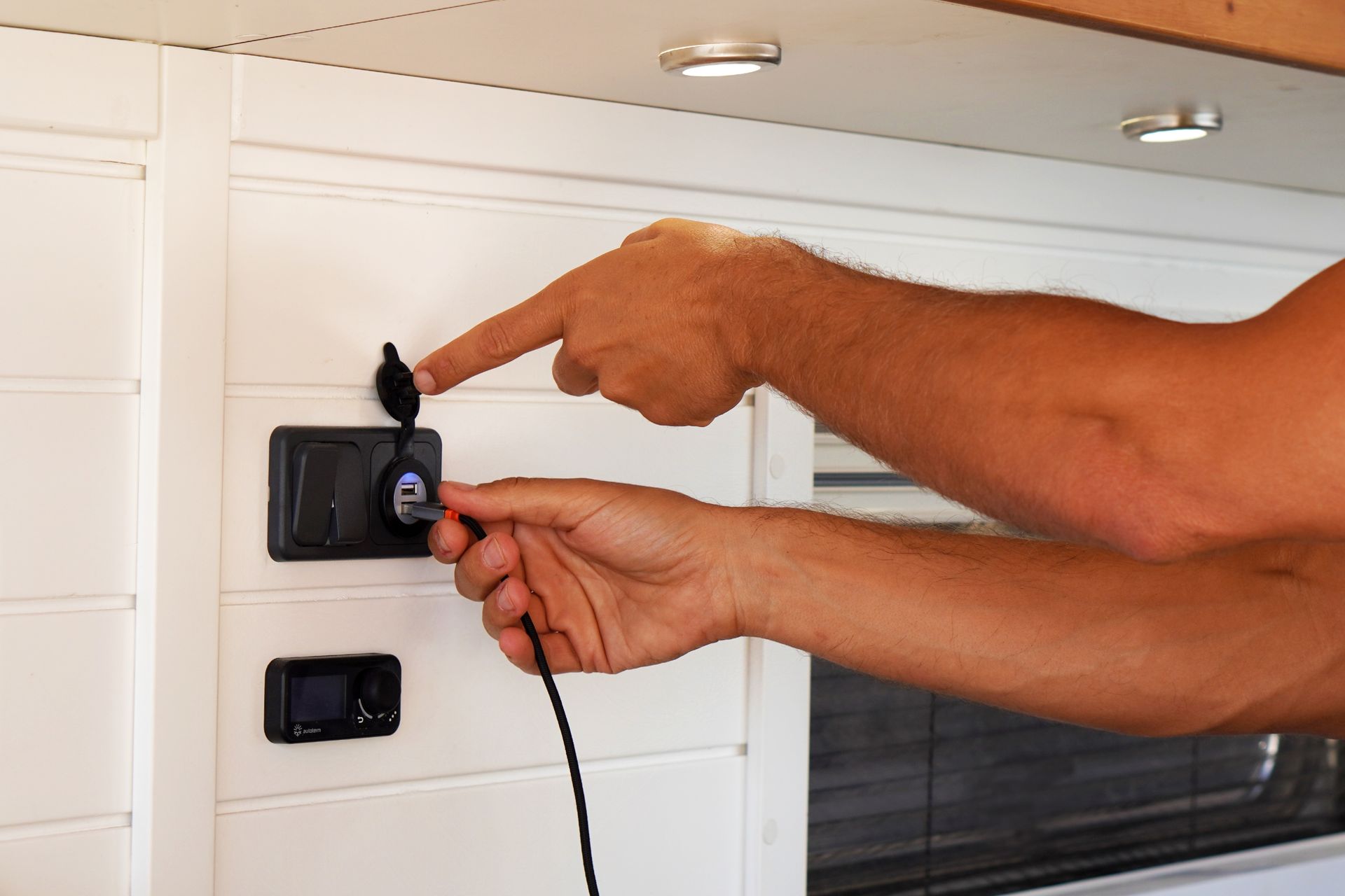 Hands plugging a cable into a black wall outlet on a white panel.