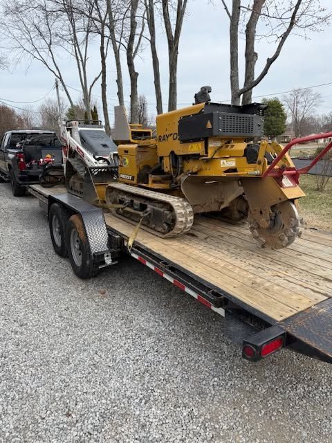 Yellow construction machine on a flatbed trailer outdoors near trees and parked vehicles.