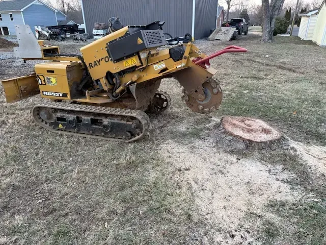 Yellow tracked trencher with a raised digging wheel parked on grass beside a concrete pad in a yard.