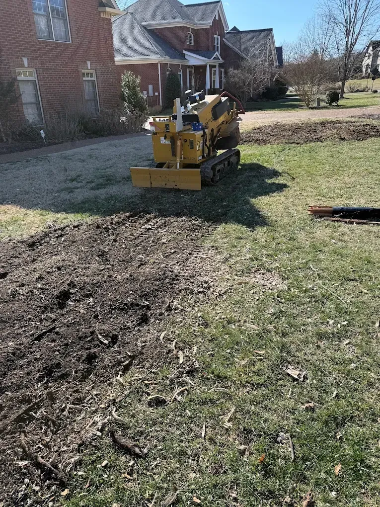 Yellow skid steer grading a dirt strip in a grassy yard beside a brick house.
