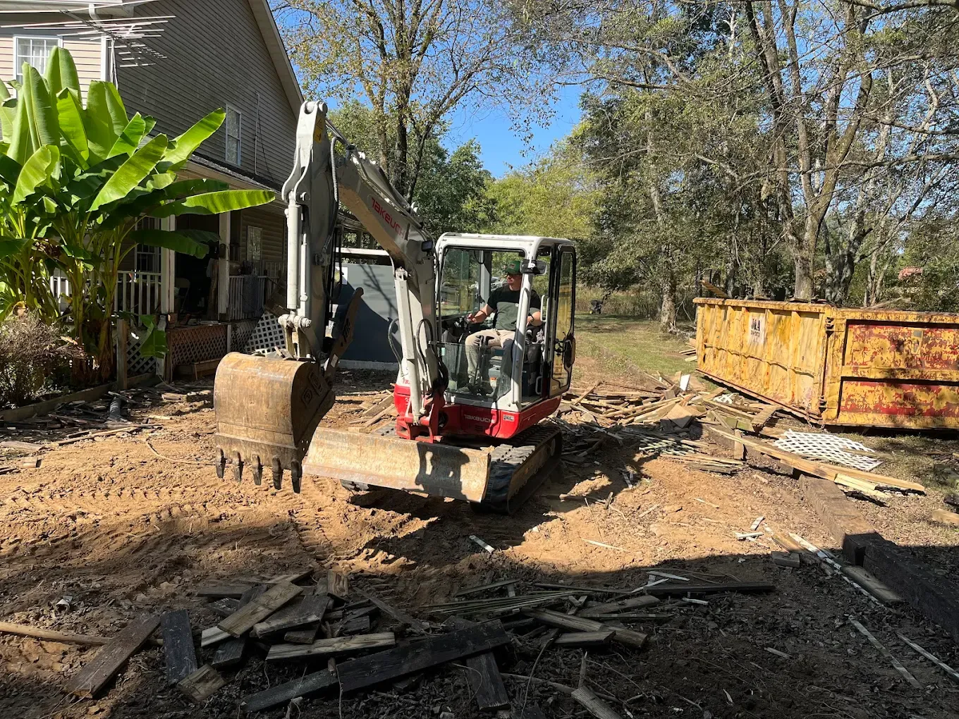 Small excavator at a dusty construction site beside a house and yellow dumpster under bright sun