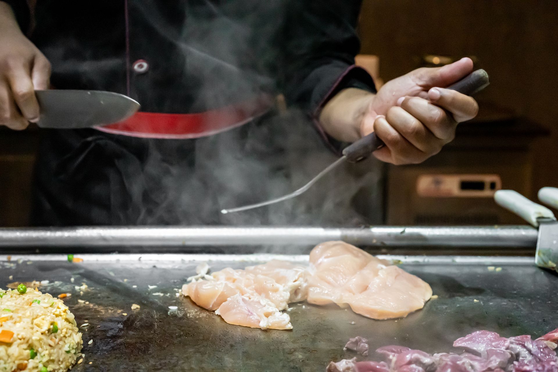 Chef cooking meat on a hot grill, holding a spatula and knife with steam rising.
