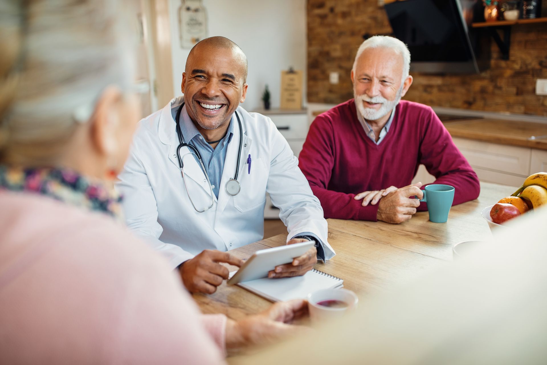 A doctor smiles while reviewing a tablet with an elderly couple in their kitchen. The man holds a mug.