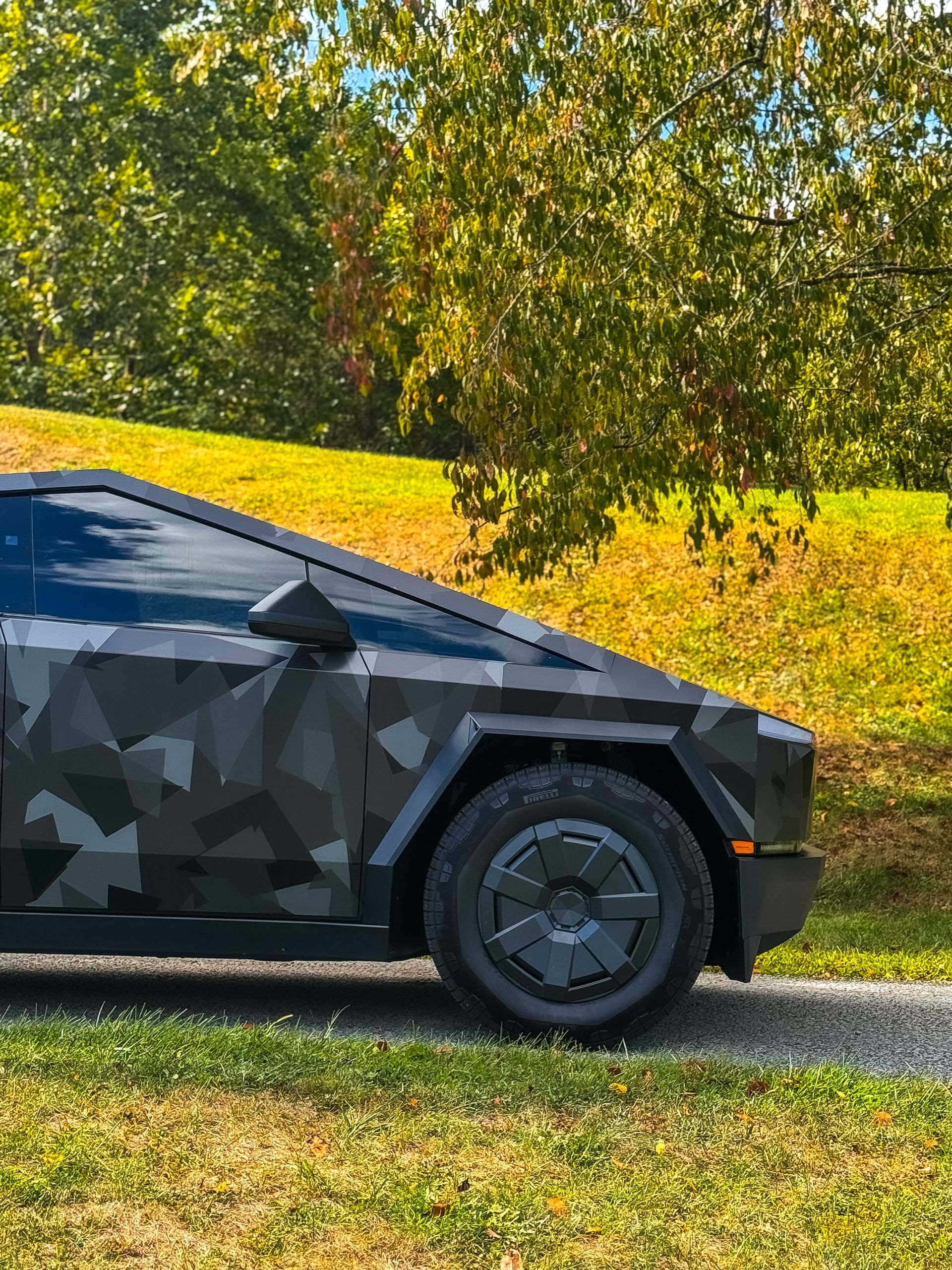 Tesla Cybertruck with dark gray camouflage wrap, parked on a path with trees in background.