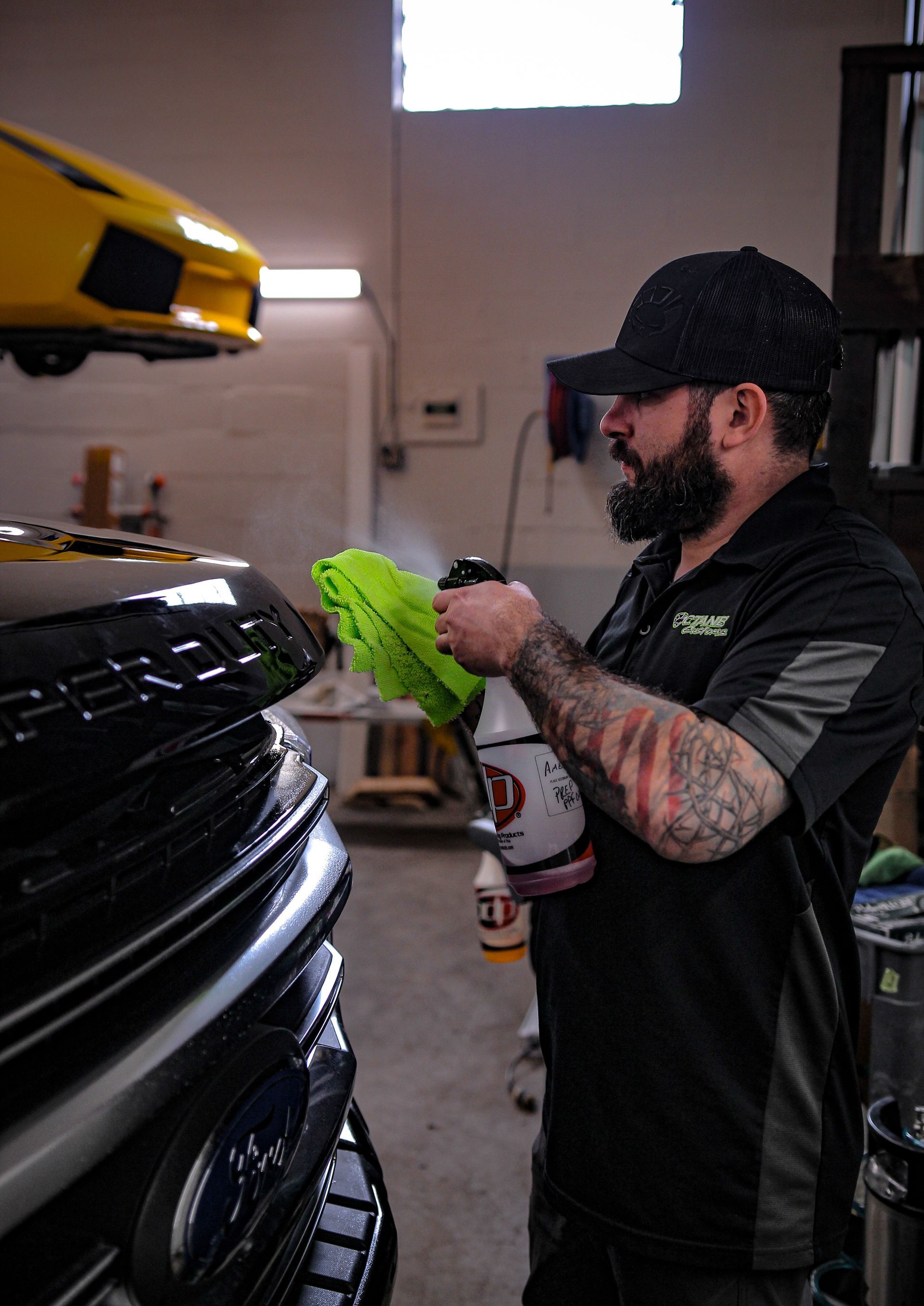 Man sprays car detailer on a black car grille, wiping with a green cloth. A yellow sports car is above.