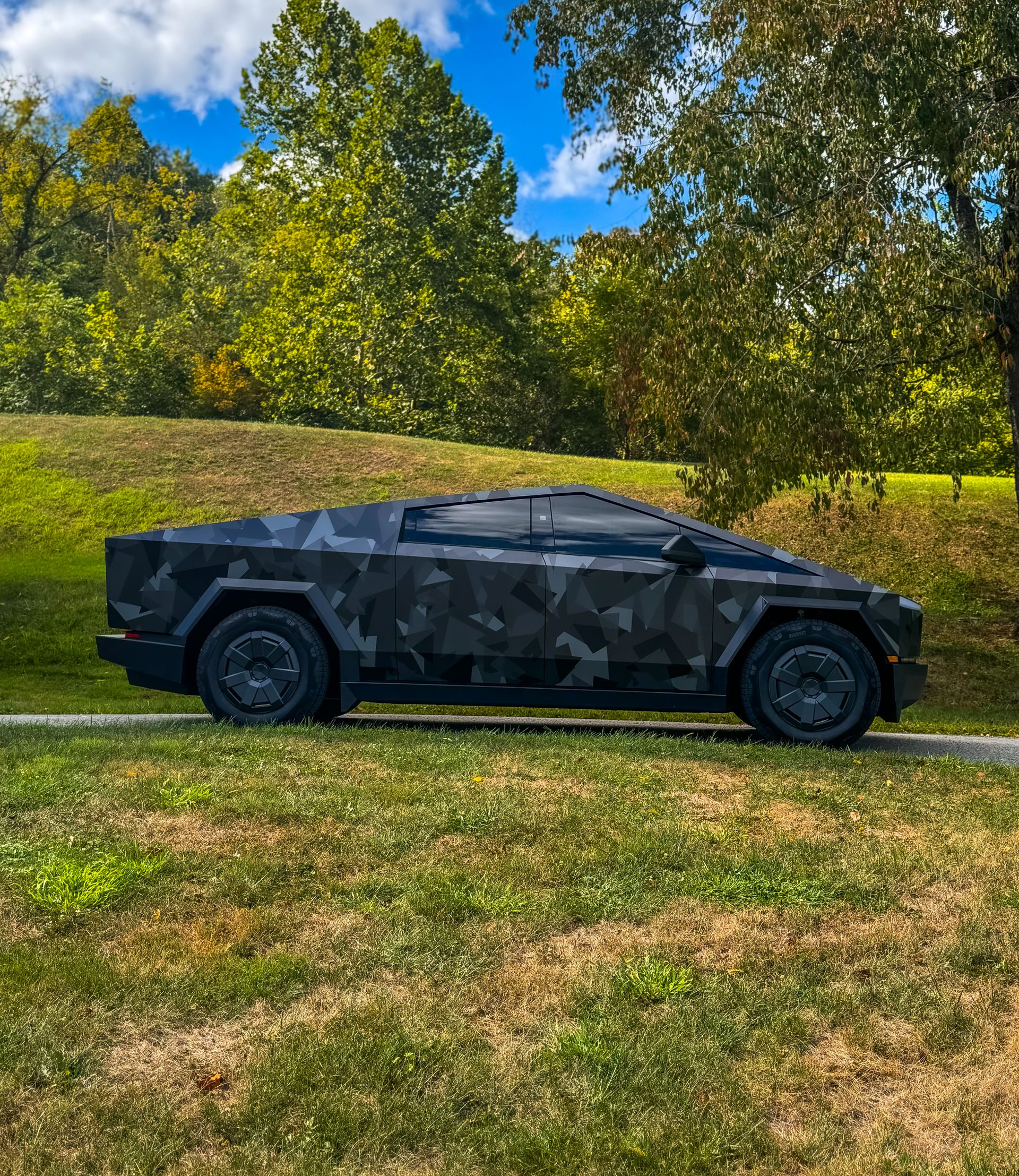 Tesla Cybertruck wrapped in black camouflage parked on grass, trees and blue sky background.