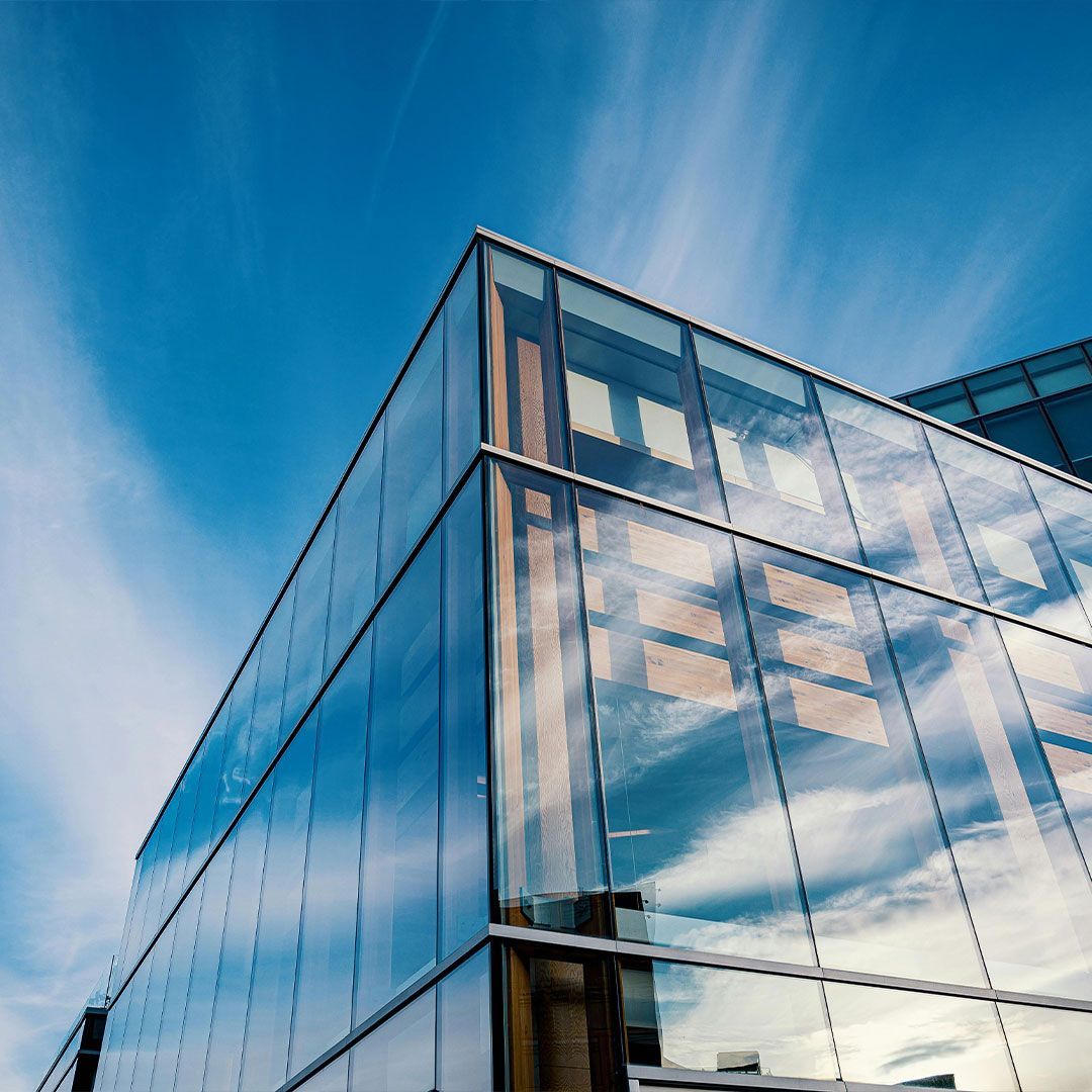 Glass-walled modern office building against a blue sky, reflecting clouds and surrounding buildings.